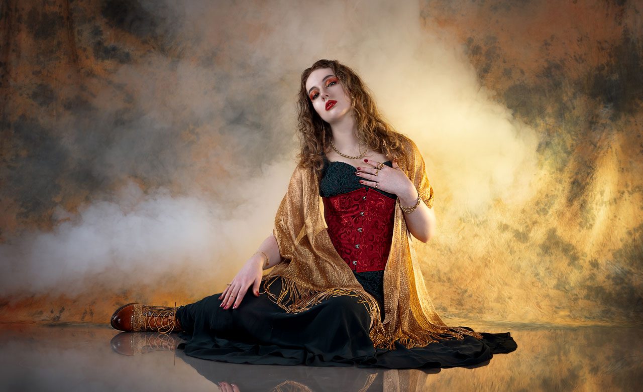 Leah seated elegantly on the floor against a gold background with soft smoke, wearing a red, gold, and black outfit.